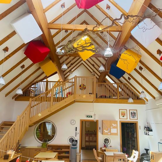 Ein Raum mit Holzbalken, einer Treppe und hängenden Dekorationen. Es gibt Tische und Stühle, ein rundes Fenster und eine Uhr an der Wand.