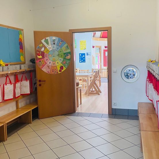 Ein Klassenzimmer mit einer Holztür und einem bunten Kalender. Taschen hängen an der Wand, und ein kreisförmiges Kunstwerk ist an der Wand. Der Raum hat weiße Fliesen, Holzbänke und eine Tür, die in einen anderen Raum mit einem Tisch und Stühlen führt.