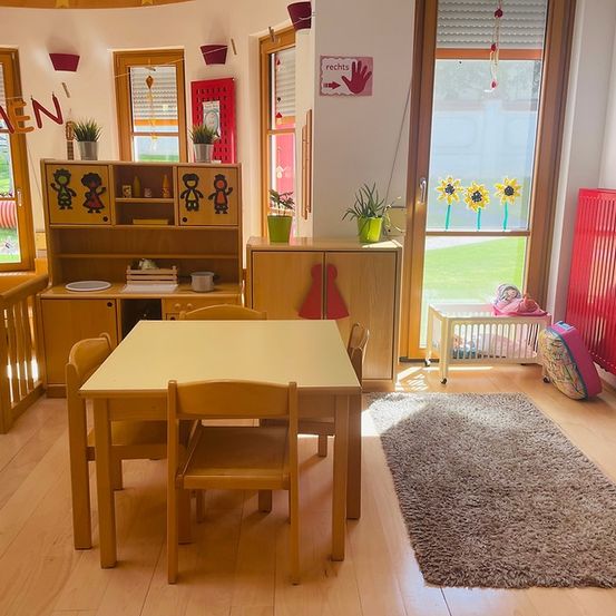 Ein Raum mit Holzmöbeln, einem Tisch und Stühlen. Auf dem Tisch stehen Topfpflanzen, ein Teller und eine Tasse. Der Raum hat Fenster, einen Heizkörper und ein Babybett.