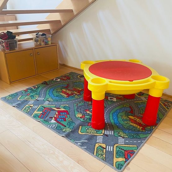 Ein Spielbereich mit einem runden gelben Tisch auf einem Teppich, der eine Straßenkarte zeigt. Ein Holzschrank mit Behältern darauf steht neben der Treppe.
