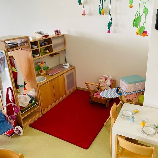 Ein Spielzimmer mit rotem Teppich, einem hölzernen Spielzeugschrank, einem Tisch mit Tellern und einem Telefon an der Wand.