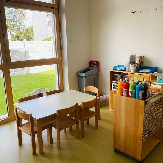 Ein Raum mit einem Tisch und Stühlen, einem Regal mit Malutensilien und einem großen Fenster mit Blick auf eine Rasenfläche.