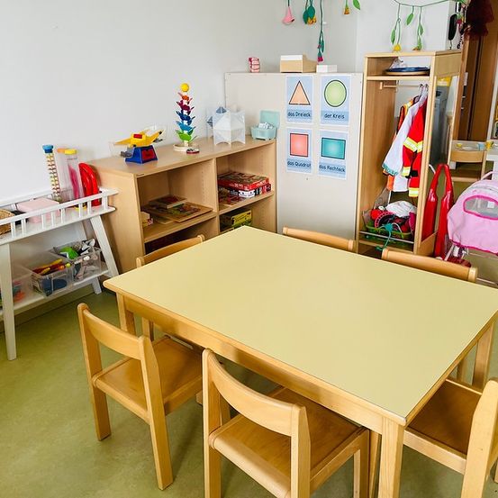 Ein Kinderspielzimmer mit einem gelben Tisch, Holzstühlen und Regalen voller Spielzeug. Der Raum hat auch einen kleinen Kühlschrank, einen Kleiderständer und einen Korb auf einem Ständer.