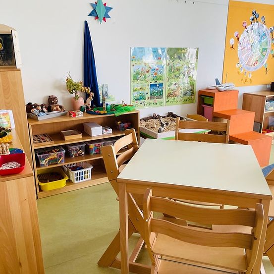 Ein Spielzimmer mit einem weißen Tisch und Stühlen. Ein Holzregal mit Spielzeug, Büchern und Aufbewahrungsboxen. Ein gelbes Wandposter und eine blaue Flagge. Ein kleines Flugzeug auf dem Regal.