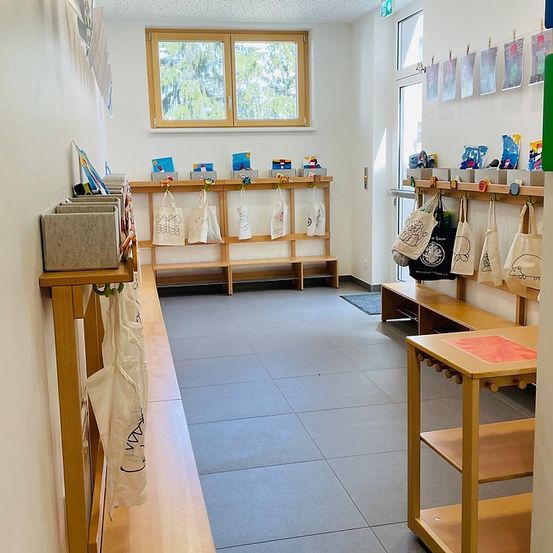 Ein leerer Klassenraum mit Holzbänken, Aufbewahrungsboxen und Taschen an Kleiderbügeln auf beiden Seiten. Der Raum hat graue Fliesenböden, weiße Wände und ein Fenster mit Blick auf Bäume.