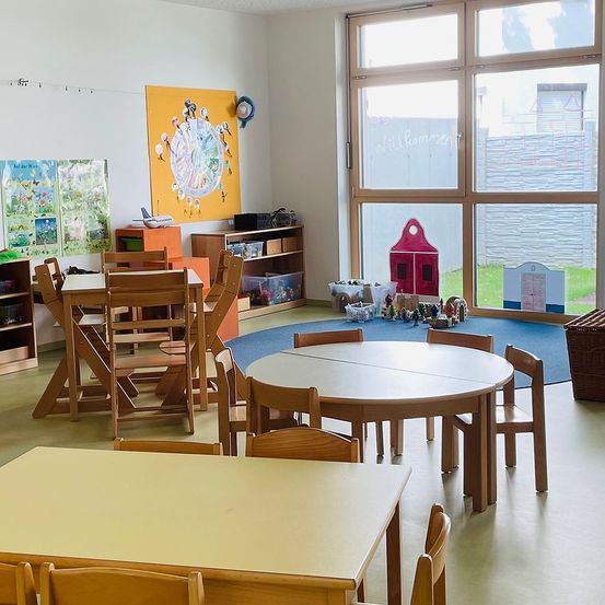 Ein Kinderzimmer mit Holztischen und Stühlen, einem runden Tisch mit Spielzeug, einem großen Fenster mit Blick nach draußen und Dekorationen an der Wand.