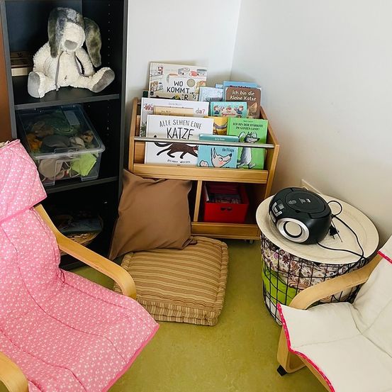 Eine gemütliche Ecke mit einem Bücherregal, einem Plüschtier, einem Kissen und einem Lautsprecher auf einem kleinen Tisch. Bücher und Spielzeug sind ordentlich aufgestellt. Ein Stuhl und eine Kissenbank bieten Sitzgelegenheiten.
