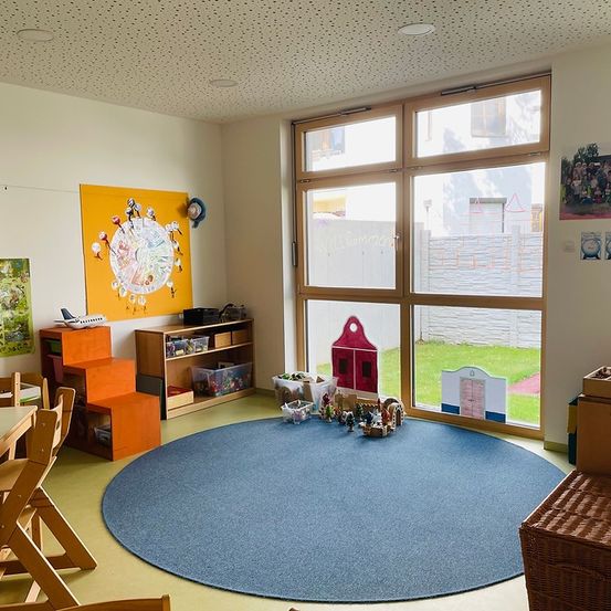 Ein gemütliches Spielzimmer mit einem großen blauen Kreisteppich. Spielzeug und Bücher sind verstreut. Ein hölzerner Tisch mit Stühlen befindet sich links. Eine gelbe Tafel mit einem Design ist an der Wand montiert. Eine Glastür öffnet sich zu einem Grasbereich.