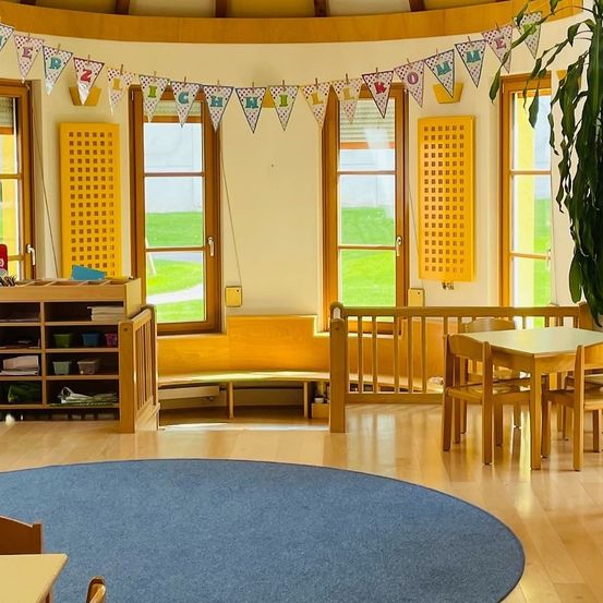 Ein Klassenzimmer mit großen Fenstern und einem runden blauen Teppich. Es gibt einen Holztisch mit Stühlen und ein Regal voller Bücher. Dekorative Wimpelketten hängen oben.