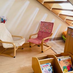 Ein gemütliches Zimmer mit zwei Holzstühlen, einer Leseecke mit Büchern und einer Treppe nach oben. Ein kleines blaues Spielzeug sitzt auf einem weißen Stuhl.