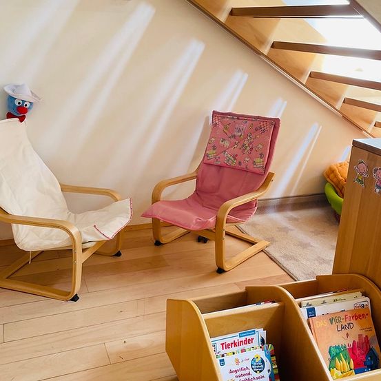 Ein gemütliches Zimmer mit zwei Holzstühlen, einer Leseecke mit Büchern und einer Treppe nach oben. Ein kleines blaues Spielzeug sitzt auf einem weißen Stuhl.