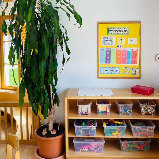 Ein Raum mit einem Holzregal und Spielzeug in durchsichtigen Plastikboxen. Eine große Pflanze und ein Wochenplan an der Wand. Eine Treppe befindet sich links.