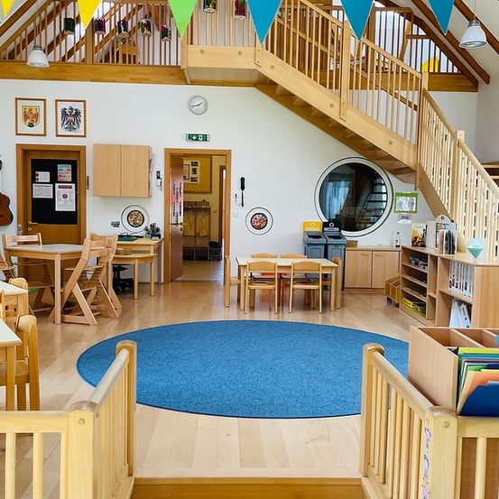 Ein holzfarbener Innenraum mit blauem Teppich, Treppe und Anschlagtafel. Der Raum hat Holzstühle und Tische, mit einer Gitarre an der Wand.