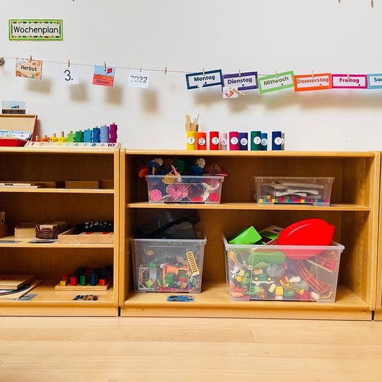 Ein Klassenzimmer mit Regalen voller verschiedener Spielzeuge und Lernmaterialien. Ein Wochenplan hängt an der Wand, und der Boden ist aus Holz.