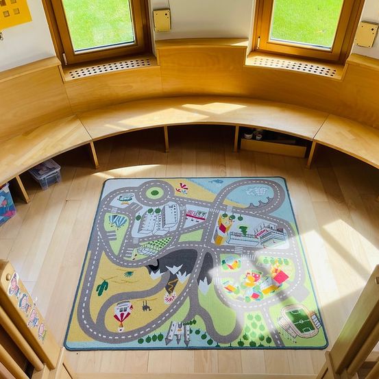 Eine Holzbank umgibt einen Spielteppich mit einer Straßenszene, Gebäuden und einem Fußballfeld. Der Raum hat große Fenster, und Sonnenlicht erhellt den Bereich.