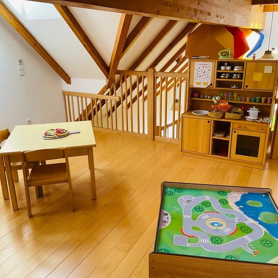 Ein Spielzimmer im Dachgeschoss mit Holzboden und Balken. Ein Tisch mit Pizza und einer Holzspielmatte. Eine hölzerne Küchenzeile und eine Treppe mit Handlauf.