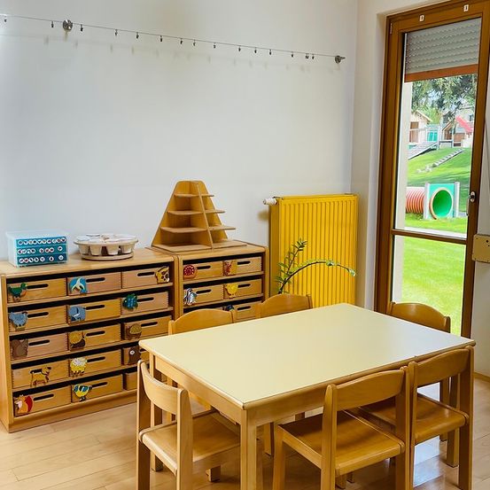 Ein Zimmer mit einem Holztisch und Stühlen. Ein Holzschrank hat Schubladen mit Tierzeichnungen. Ein Holzdreieck steht auf dem Schrank. Ein Heizkörper ist an der Wand. Eine Pflanze steht in der Nähe der Tür. Draußen ist ein Rasenbereich mit einem Tunnel.
