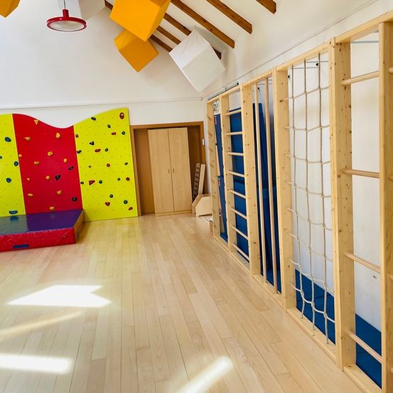 Ein großer Innenspielraum mit Holzboden, einer Holzkletterwand mit roten und gelben Abschnitten, einem Holzschrank und einer Seilleiter auf der rechten Seite.