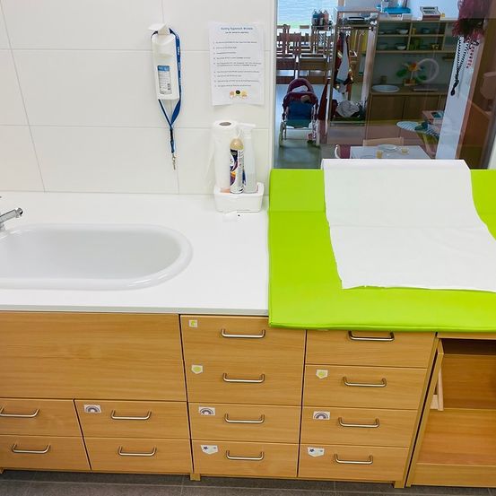 Ein Badezimmer mit einem weißen Waschbecken, Holzschubladen und einer grünen Matte. Ein Händedesinfektionsmittel und eine Flasche sind auf dem Tisch. Ein Poster mit Richtlinien ist an der Wand. Ein Spiegelbild eines Kinderwagens und Stühle ist auf der Glastür.
