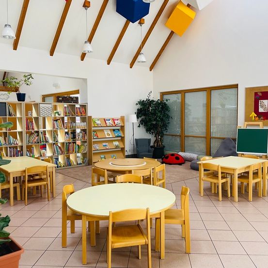 Ein heller, luftiger Raum mit Holztischen und Stühlen, einem Bücherregal voller Bücher und einer Pflanze in einem Topf. Der Raum hat einen gefliesten Boden und eine Decke mit hängenden Lampen.