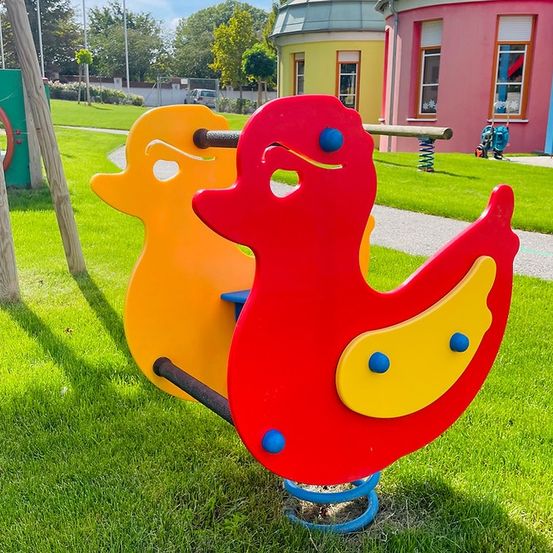 Ein bunter Spielplatz mit einem roten und einem gelben Entenspielzeug an einem sonnigen Tag. Dahinter befindet sich ein rosa Gebäude mit Fenstern.