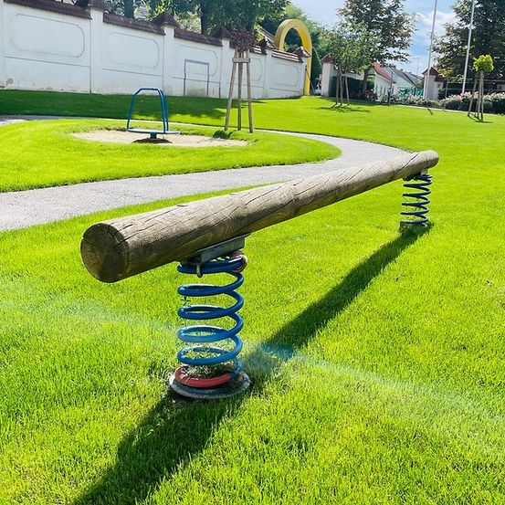 Eine Wippe auf Federn steht auf einer Rasenfläche eines Parks. Sie besteht aus einem Holzbalken. Im Hintergrund befindet sich ein Spielplatz mit Schaukeln und einer Mauer.