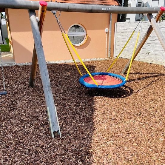 Ein Spielplatz mit einer Schaukel und einem runden Reifenschaukel unter einem Haus. Die Reifenschaukel ist blau und rot.
