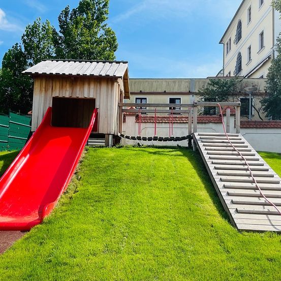 Ein Außenspielbereich mit einem roten Rutsch, Holzstufen, einem Holzhaus und einem Gebäude mit roten Ziegeln im Hintergrund.