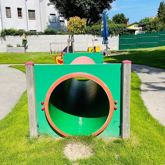 Ein grüner Tunnel mit roten Knöpfen auf einem Rasen in der Nähe eines weißen Gebäudes mit Pflanzen.