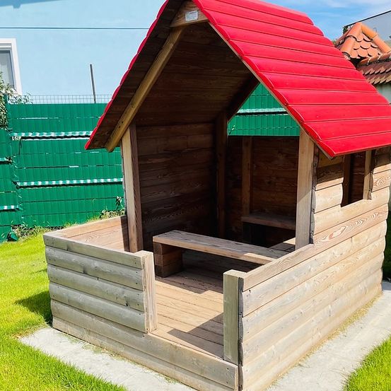 Ein Holzspielhaus mit rotem Dach befindet sich auf einem Rasenbereich. Das Spielhaus verfügt über eine Bank im Inneren und eine kleine Holztür.