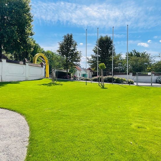 Ein großer, gut gepflegter Rasen mit einer weißen Mauer und einem gelben Bogen. Bäume und Pflanzen sind über das Gelände verstreut. Ein Auto ist in einem umzäunten Bereich geparkt.