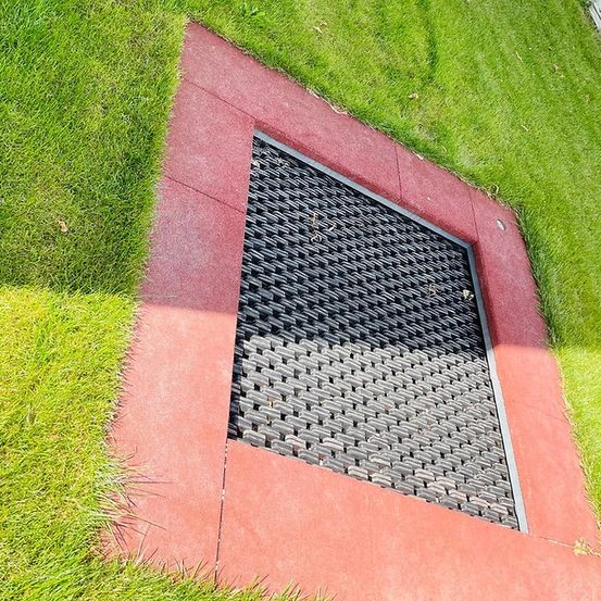 Ein rotes quadratisches Podest mit einer quadratischen schwarzen Gitterroste in der Mitte, umgeben von grünem Gras.