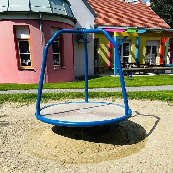 Ein Spielplatz mit einer blauen Drehscheibe in einer Sandgrube. Dahinter befindet sich ein rosa Gebäude mit Fenstern und ein gelbes Gebäude mit einer Terrasse.