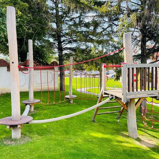 Ein Außenspielplatz mit Seilleitern und einer Kletterwand, umgeben von Bäumen und grünem Rasen.