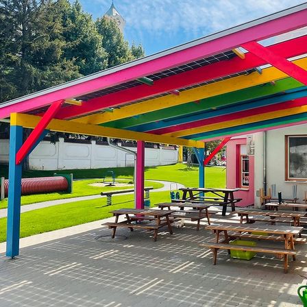 Ein farbenfroher Pavillon mit Solarpanelen und Holzpicknicktischen unter einem lebhaften Dach. Ein Spielplatz und eine grüne Wiese im Hintergrund.