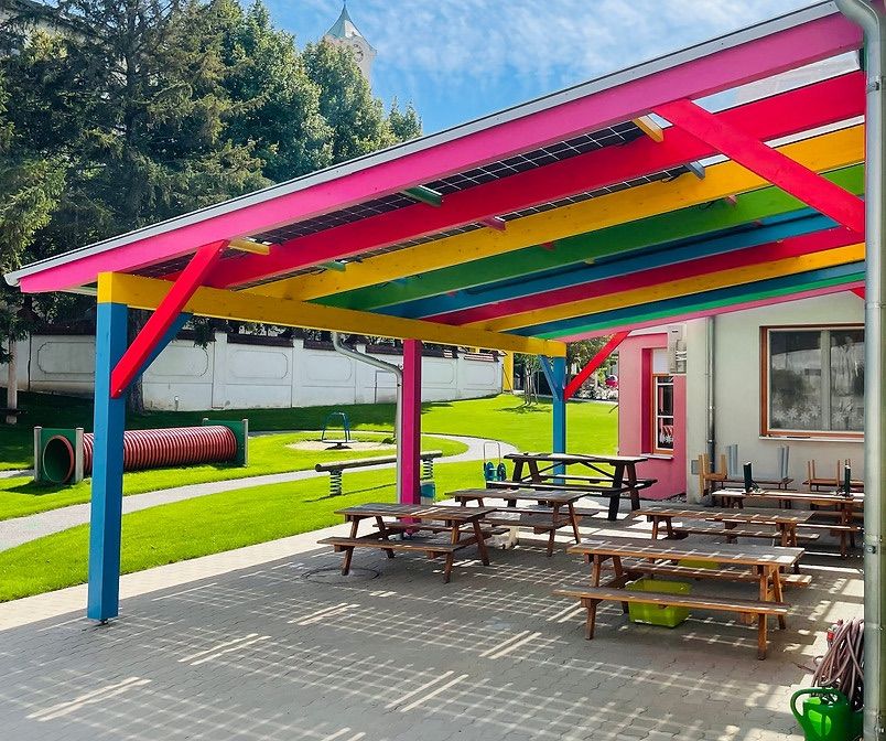 Ein farbenfroher Pavillon mit Solarpanelen und Holzpicknicktischen unter einem lebhaften Dach. Ein Spielplatz und eine grüne Wiese im Hintergrund.