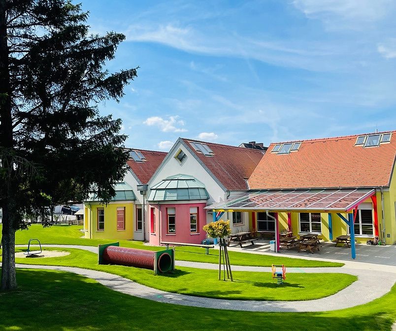 Ein leuchtendes, farbenfrohes Haus mit einem grünen Rasen und einem Spielplatz. Ein Baum steht neben dem Haus. Unter einem Vordach befinden sich Bänke und Picknicktische.