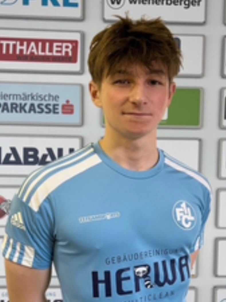 Ein junger Mann in einem blauen Fußballtrikot mit weißen Streifen und einem Logo auf der Brust steht vor einer Wand mit verschiedenen Schildern.