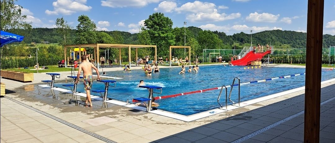 Ein großer Außenpool mit vielen schwimmenden und spielenden Menschen. Eine rote Rutsche befindet sich in der Nähe des Pools, und es gibt Bänke und einen Spielplatz.