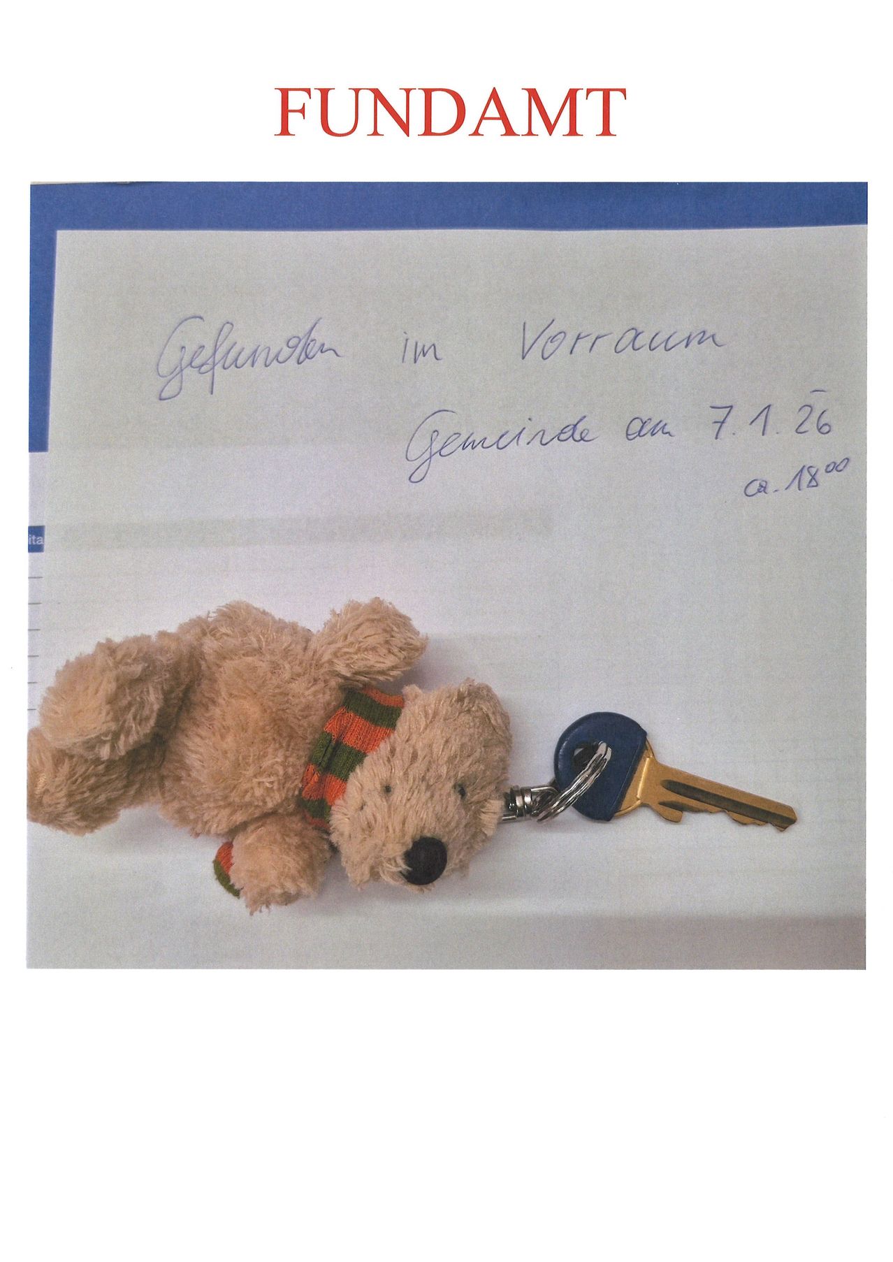 Ein gefundener brauner Teddybär mit einem blauen Schlüssel auf einem weißen Papier mit blauen Rändern. Das Papier hat handgeschriebenen Text 'Gefunden im Vorraum Gemeinde am 7.1.26 q. 18:00'.