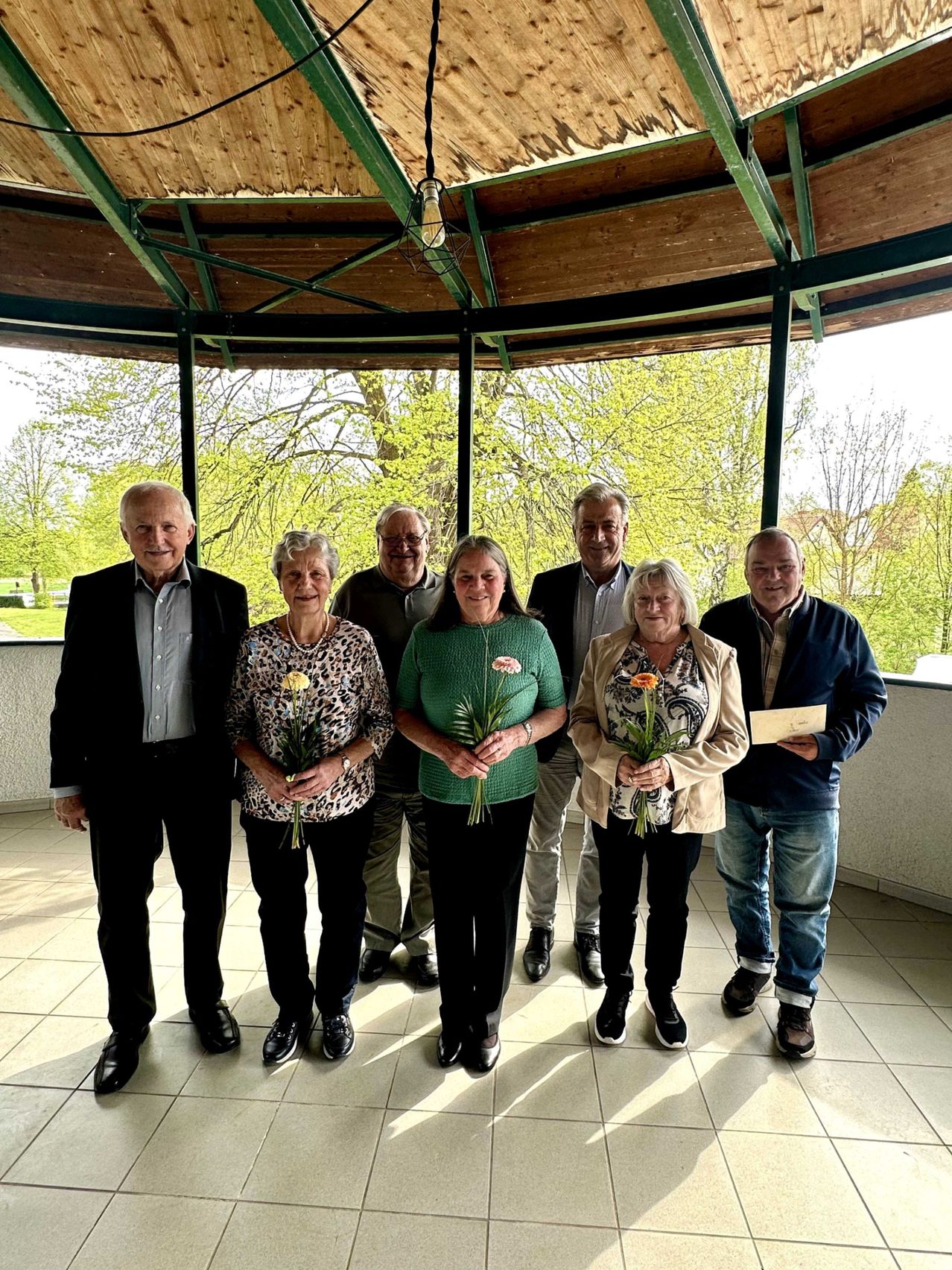 Eine Gruppe von Menschen, wahrscheinlich eine Familie, steht zusammen in einem Pavillon, lächelt und posiert für ein Foto. Sie halten Blumen in den Händen und scheinen einen Anlass zu feiern. Der Pavillon ist von einem Rasen mit Bäumen umgeben.