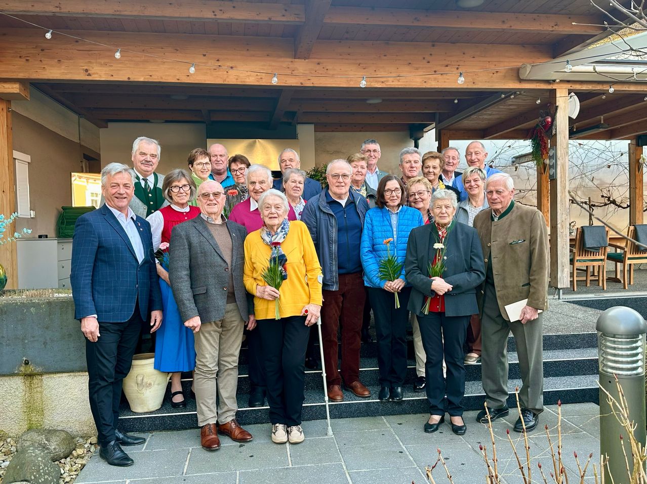 Eine Gruppe von Menschen steht auf einer Veranda, einige halten Blumen. Sie scheinen für ein Foto zu posieren. Die Veranda hat eine Holzdecke und Geländer.