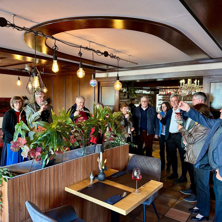 Eine Gruppe von Menschen versammelt sich in einem Restaurant und hebt ihre Gläser zum Anstoßen. Der Raum hat Holzwände, einen Holztisch mit Weingläsern und Pflanzen. Es gibt hängende Lampen und eine Kristallleuchte.