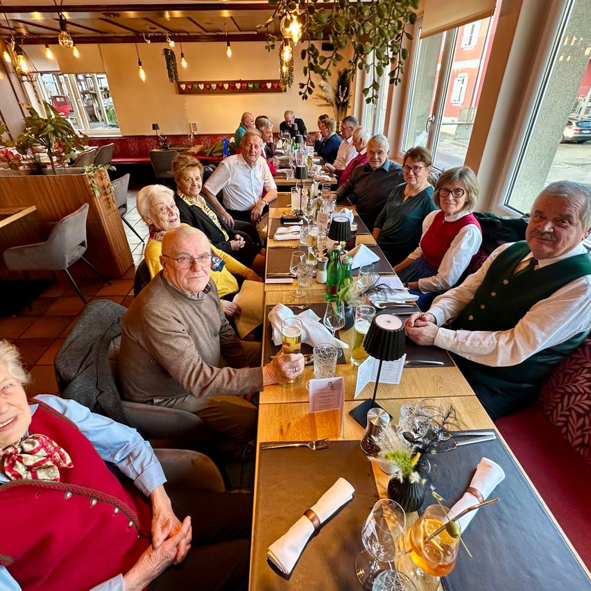 Eine Gruppe älterer Menschen sitzt an einem langen Tisch in einem Restaurant, lächelt und posiert für ein Foto. Der Tisch ist mit Getränken, Gläsern und Servietten gefüllt. Das Restaurant hat große Fenster und Topfpflanzen.