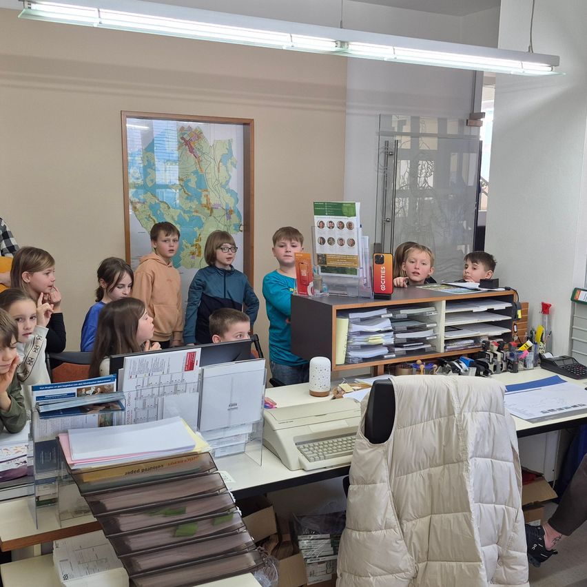 Kinder stehen in einem Klassenzimmer und blicken auf einen Schreibtisch mit Computer, Büchern und einer Karte an der Wand. Sie wirken interessiert und aufmerksam.