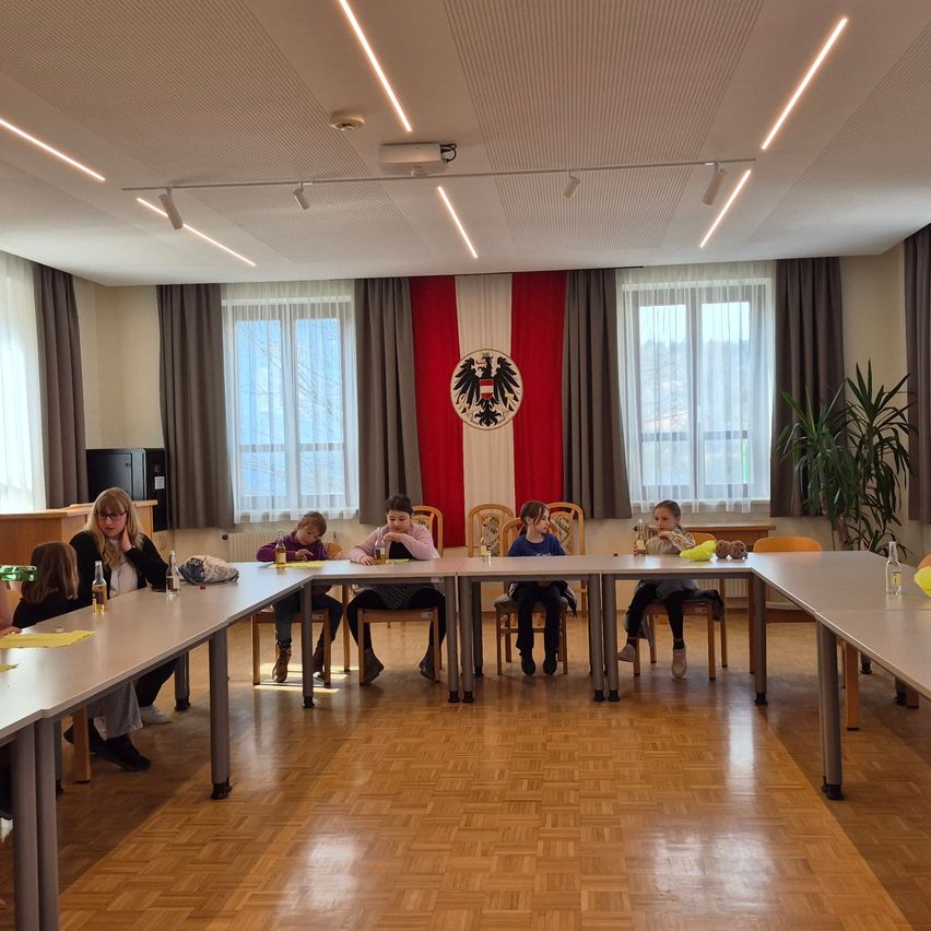 Eine Gruppe von Kindern und einer Frau sitzen an einem langen Tisch in einem Raum mit Holzboden und großen Fenstern. Hinter ihnen befindet sich eine rot-weiße Wand mit einem deutschen Wappen.