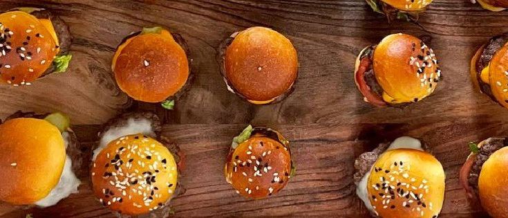 Ein Holzbrett zeigt mehrere Hamburger mit Sesamkörnern. Einige haben Salat und Käse. Die Präsentation ist ordentlich, mit in zwei Hälften geschnittenen Brötchen und sichtbaren Zutaten.