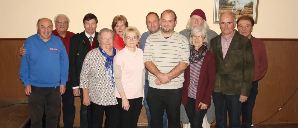 Eine Gruppe von Erwachsenen, möglicherweise Familie oder Kollegen, steht für ein Foto in einem Raum mit weißer Wand, Holzboden und gerahmtem Bild an der Wand.