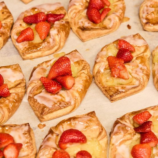 Mehrere herzförmige Gebäckstücke mit Erdbeerscheiben und Glasur sind auf einem Backblech angeordnet. Jedes Gebäck ist mit einer gelben Creme gefüllt und mit Erdbeeren und Glasur belegt.