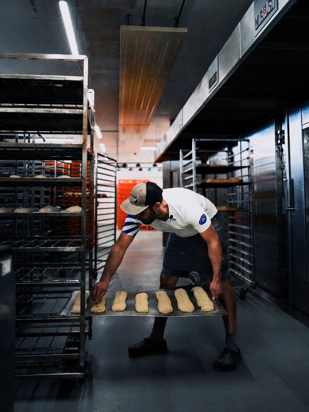 Ein Mann arbeitet in einer Bäckerei. Er trägt eine Kappe, ein weißes Shirt und einen schwarzen Rock. Er hält eine Ablage mit Teig darauf. Dahinter befinden sich Regale mit Böden, auf denen Brot liegt.